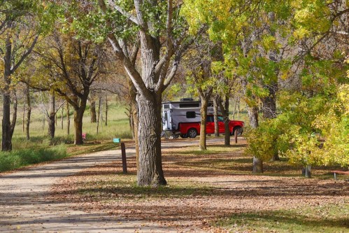 A red camper parked among trees with autumn leaves, along a dirt path in a serene outdoor setting.