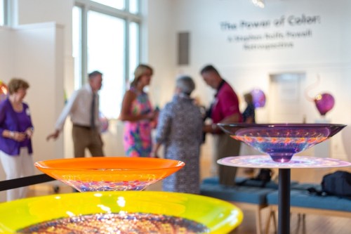 Colorful glass art pieces in the foreground with people mingling at an art exhibit in a bright, spacious gallery.