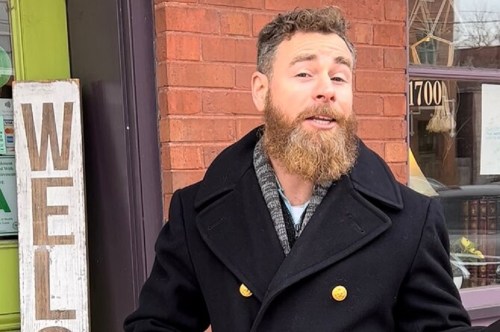 A man with a beard stands outside a shop, smiling and wearing a black coat, next to a wooden "Welcome" sign.