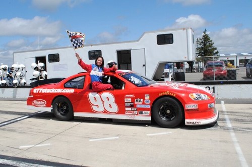 A red race car with the number 98, featuring a driver holding a checkered flag, parked on a racetrack.