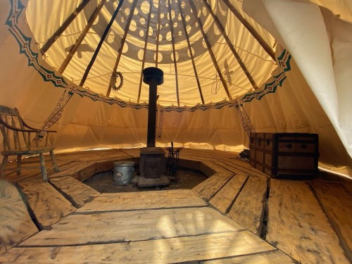 Interior of a tent with wooden flooring, a stove in the center, and a chair, illuminated by natural light.