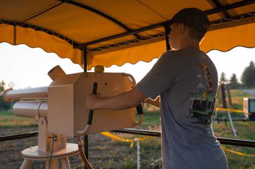 A young person operates a telescope under a yellow canopy, with a scenic outdoor background.