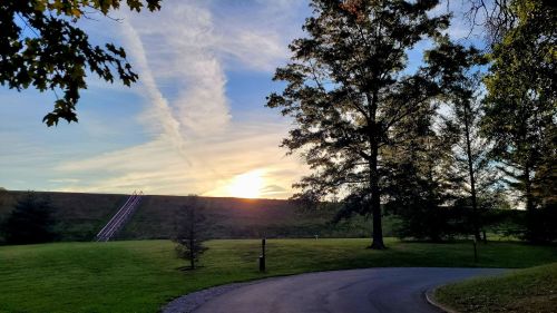 A serene landscape at sunset, featuring trees, a grassy area, and a winding road leading towards a hill.