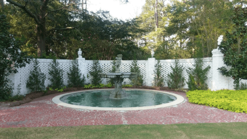 A serene garden scene featuring a circular fountain surrounded by greenery and a white brick wall.