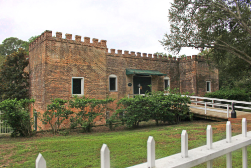Historic brick building with a castle-like design, surrounded by greenery and a white picket fence.