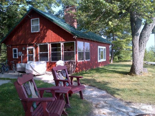 A red cabin with a green roof surrounded by trees, featuring two red chairs in front and a gravel path.