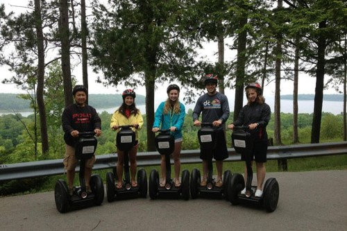 Five people on Segways in a forested area, smiling and wearing helmets, with a scenic view in the background.