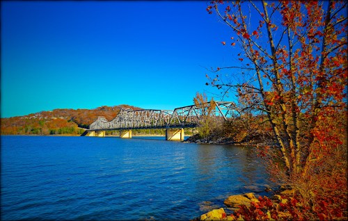 A metal bridge spans a calm blue lake, surrounded by autumn foliage and hills under a clear blue sky.