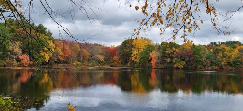A serene lake surrounded by vibrant autumn foliage under a cloudy sky. Reflections of trees shimmer on the water's surface.
