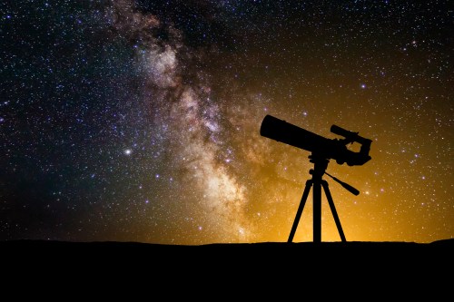 A silhouette of a telescope against a starry night sky with the Milky Way and a warm glow in the background.