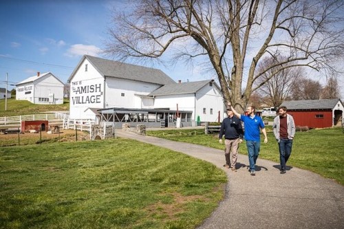 Three men walk along a path in an Amish village, with a large barn and green fields in the background.