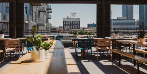 A rooftop dining area with wooden tables and plants, overlooking a city skyline with buildings in the background.