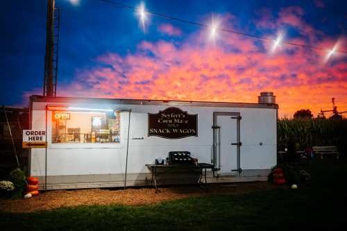 A snack wagon with an "Open" sign, set against a colorful sunset and string lights in the background.