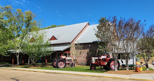 A rustic building with a metal roof, surrounded by trees and two vintage tractors in front on a sunny day.