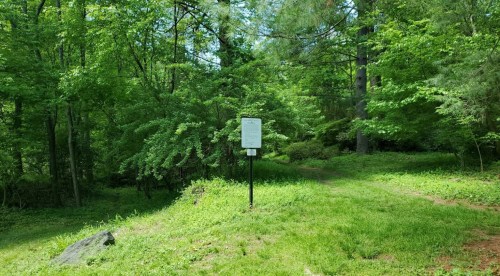 A sign stands on a grassy path surrounded by lush green trees in a wooded area.