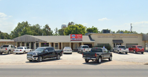 A building with a sign, surrounded by parked cars, under a clear blue sky. Trees are visible in the background.