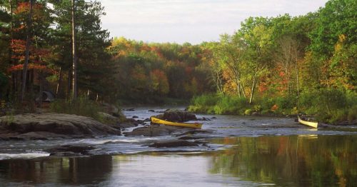 A serene river scene with colorful autumn trees and two yellow canoes on the water, surrounded by rocky banks.