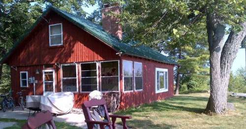 A red cabin with a green roof surrounded by trees, featuring a porch and outdoor seating in a grassy area.