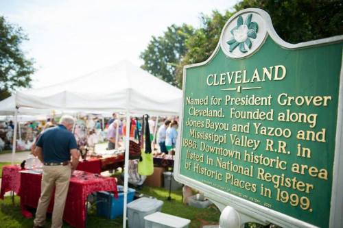 A green historical marker for Cleveland, with a busy outdoor market in the background under white tents.