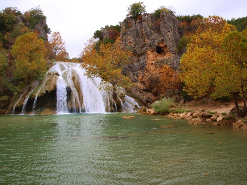 A serene waterfall cascades into a calm, green pool, surrounded by autumn-colored trees and rocky cliffs.