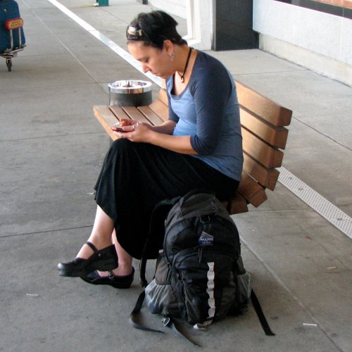 A woman sitting on a bench, focused on her phone, with a backpack beside her in a public space.