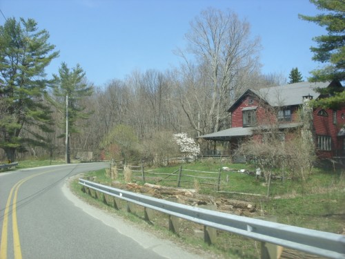 A winding road beside a red house, surrounded by trees and greenery under a clear blue sky.