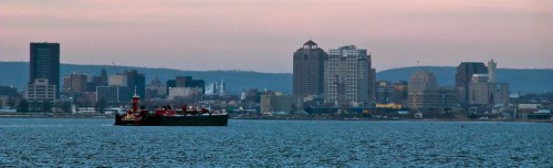 A cargo ship sails in the foreground with a city skyline in the background during twilight.