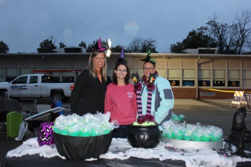 Three women in festive hats stand behind a table with green treats and a cauldron, set against a school backdrop.