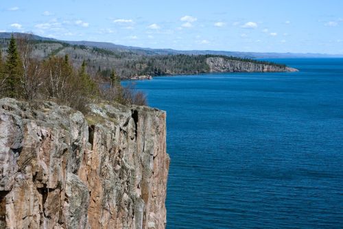 A rocky cliff overlooks a calm blue lake, surrounded by lush green hills and a clear sky with scattered clouds.