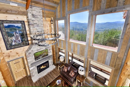 Cozy cabin interior with a stone fireplace, large windows, and mountain views, featuring wooden walls and rustic decor.