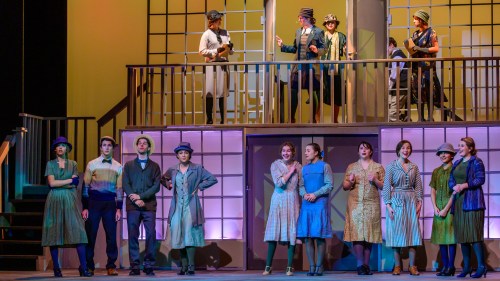 A theater performance featuring a diverse cast in 1920s costumes, with some actors on a balcony and others on stage.