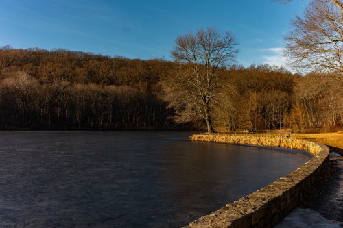A serene lake surrounded by trees, with a stone path along the water's edge under a clear blue sky.
