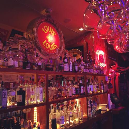 A dimly lit bar with a wooden shelf displaying various bottles of liquor and colorful lights in the background.