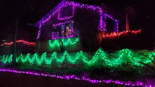 A house decorated with colorful purple and green lights, illuminating the exterior and surrounding landscape at night.