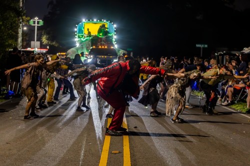 A performer in a red suit dances in the street, surrounded by dancers in costumes during a nighttime parade.