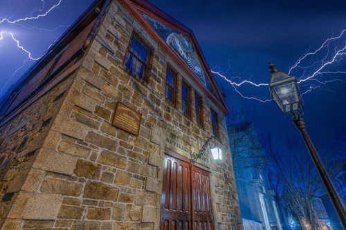A historic stone building illuminated by lightning in a dark, stormy sky, with a streetlamp in the foreground.