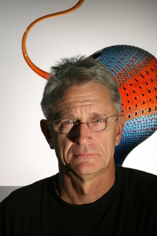 A man with glasses and gray hair stands in front of a colorful abstract background.