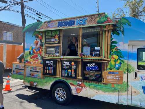 A colorful Kona Ice truck with a tropical theme, featuring a menu of flavors and a staff member serving customers.