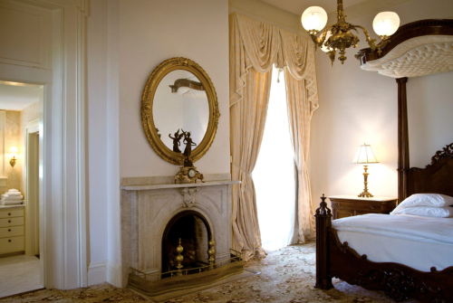 Elegant bedroom featuring a vintage bed, ornate mirror, fireplace, and draped windows with soft lighting.
