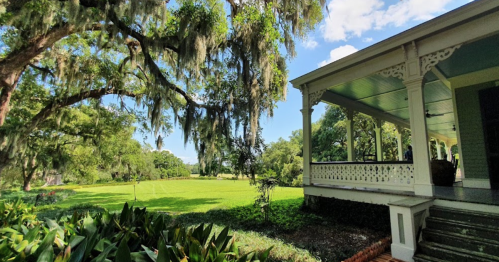 A sunny landscape featuring a green lawn, trees with Spanish moss, and a porch with decorative railings.