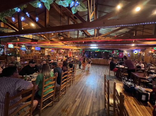 A lively restaurant interior with wooden decor, colorful lights, and diners enjoying their meals at long tables.