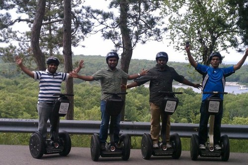 Four people on Segways, smiling and posing with arms raised, surrounded by trees and a scenic landscape.
