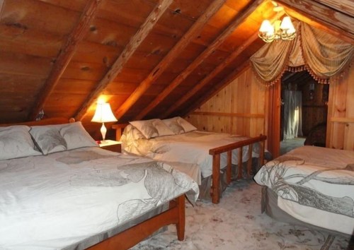 Cozy attic bedroom with two beds, wooden walls, and a warm lamp, creating a rustic and inviting atmosphere.