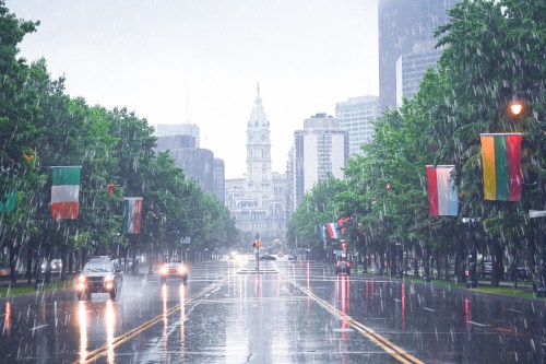 A rainy city street lined with trees and flags, featuring a view of a tall building in the background.