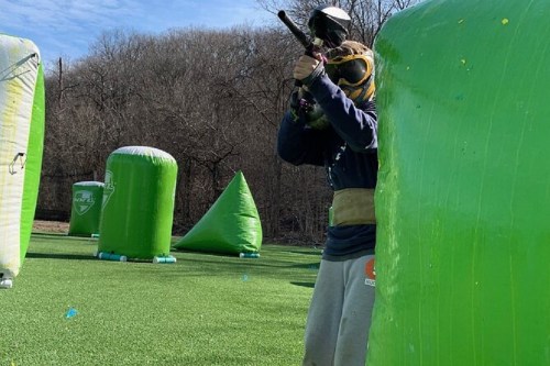A person in protective gear aims a paintball gun from behind a green inflatable barrier on a field.