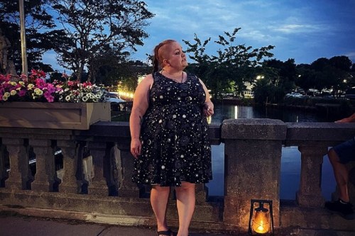 A woman in a black dress stands by a stone railing near a river, with flowers and a lantern in the evening light.