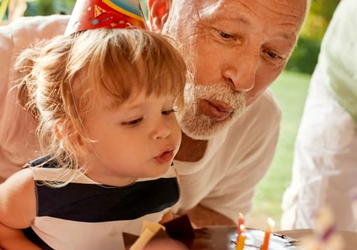 A young girl and an older man blow out birthday candles together, both wearing party hats and smiling.