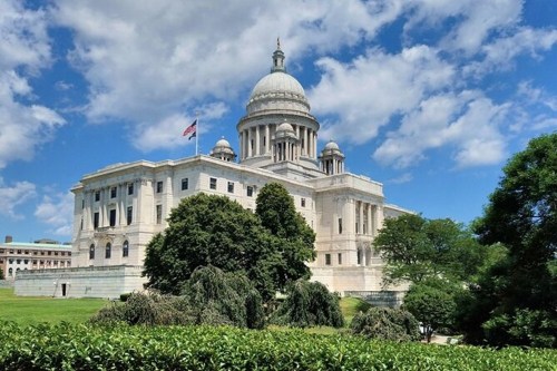 The Rhode Island State House, featuring a large dome, surrounded by greenery and a blue sky with clouds.