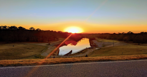 A serene sunset over a calm lake, reflecting vibrant orange and yellow hues, surrounded by trees and grassy banks.