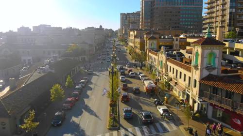 Aerial view of a busy street lined with shops and cars, bathed in sunlight, with buildings in the background.